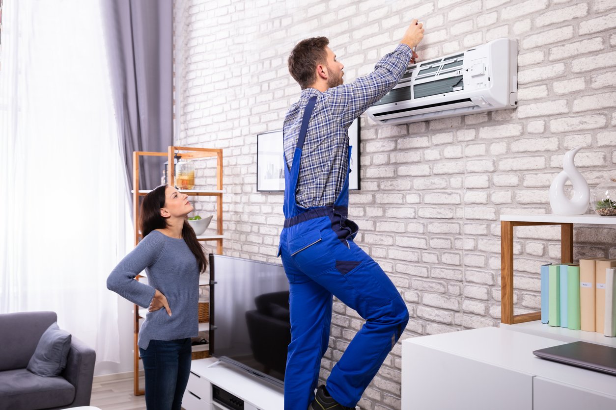 Man fixes air conditioner while woman watches.